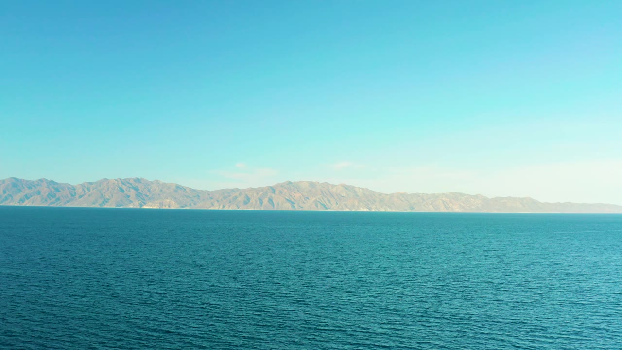 A wide sea surface with small ripples leads to a distant mountain ridge and pale blue sky in El Sargento La Ventana, Mexico, with no visible people.