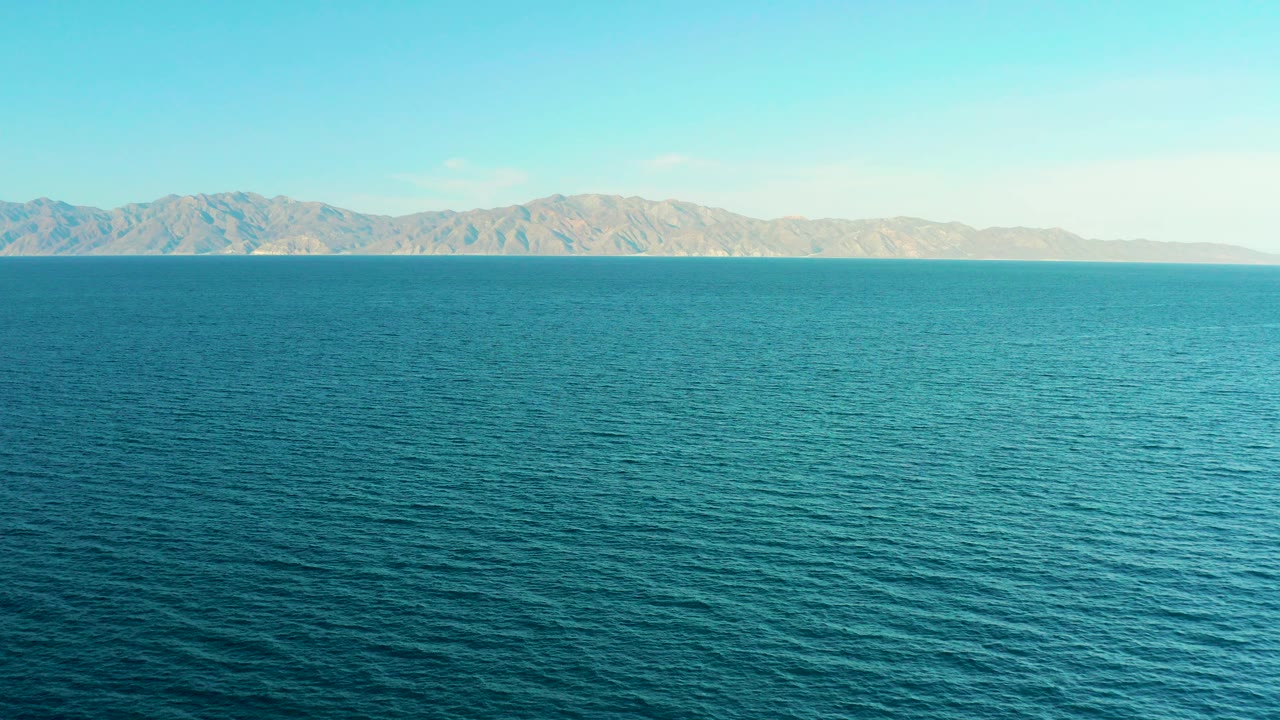 Wide view over blue-green water toward a tan mountain shoreline near El Sargento La Ventana, Mexico, with pale sky above and no visible people.