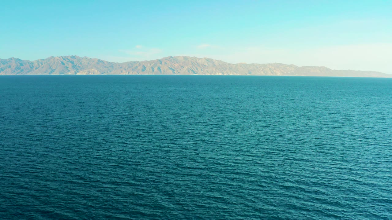 Blue sea surface extends toward low, dry mountains under a pale clear sky in El Sargento La Ventana, Mexico, with no people, vessels, or structures visible.