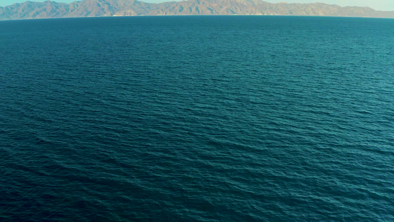 Wide aerial view over deep blue water near El Sargento La Ventana, Mexico, with tan mountains across the horizon and no visible people.