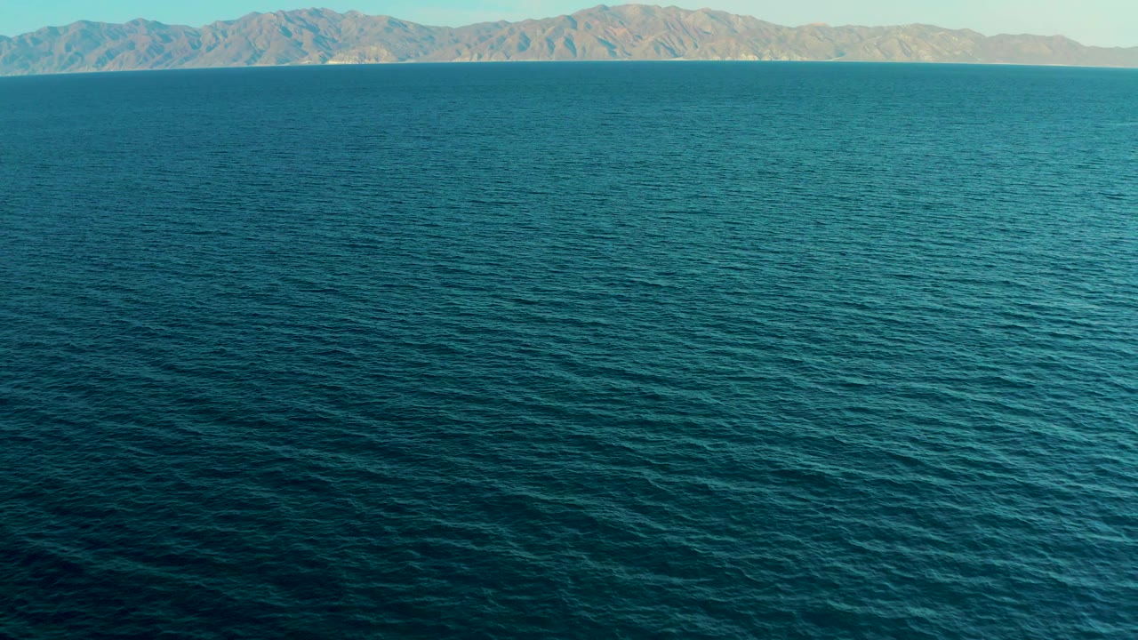 A high view shows calm teal-blue sea below distant mountains near El Sargento La Ventana, Mexico, with no visible people.