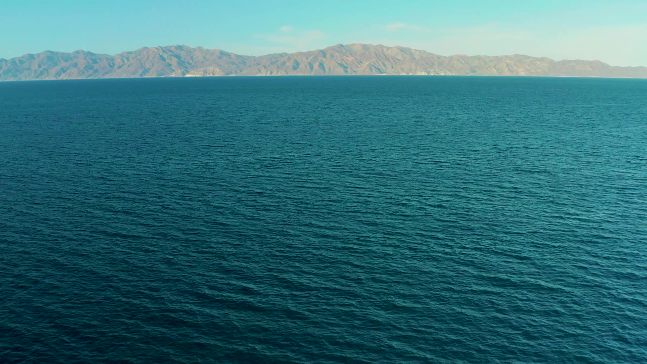 At 0:02, textured blue water fills most of the frame, leading to tan mountains beneath a clear sky in El Sargento La Ventana, Mexico, with no visible people.