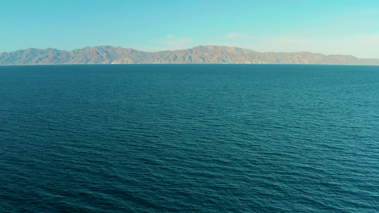 Early frame of El Sargento La Ventana, Mexico, showing blue-green ocean texture filling most of the view, pale mountains across the upper horizon, clear sky, and no visible people.