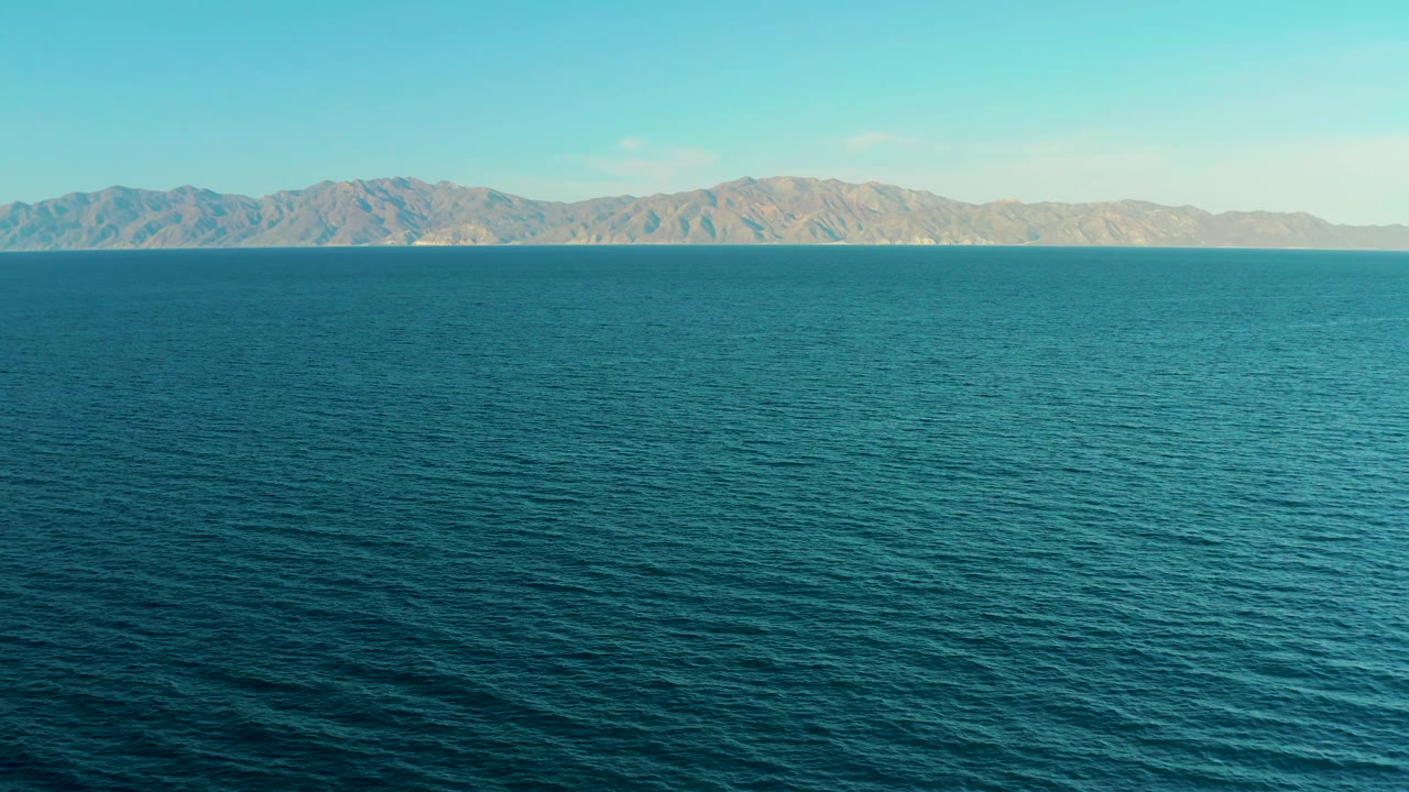 Aerial frame from El Sargento La Ventana, Mexico, with turquoise sea in the foreground, rugged brown mountains stretching across the distance, bright daylight, and no visible people.