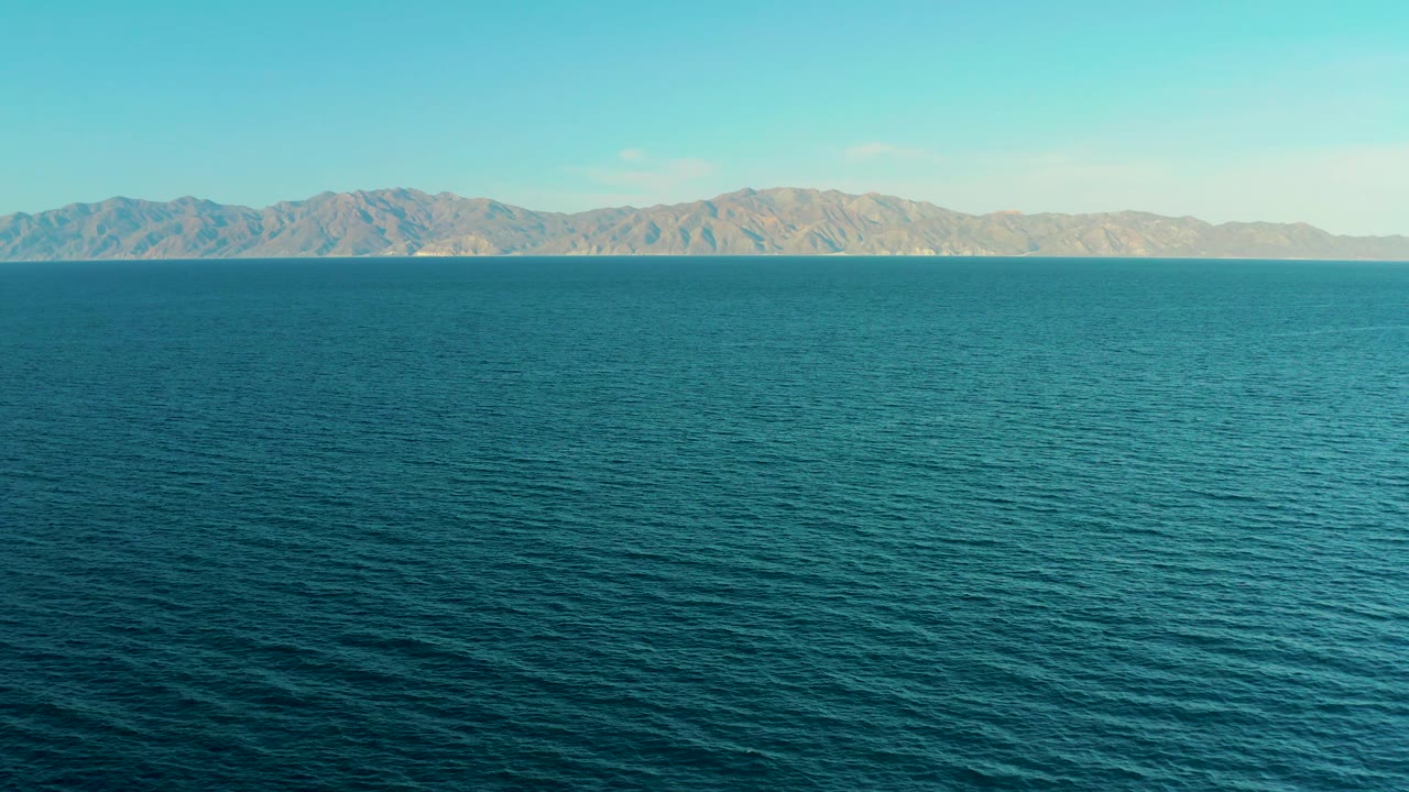 Mid-clip frame in El Sargento La Ventana, Mexico, with a calm ocean expanse below a pale mountain ridge, open blue sky, a straight horizon, and no visible people.
