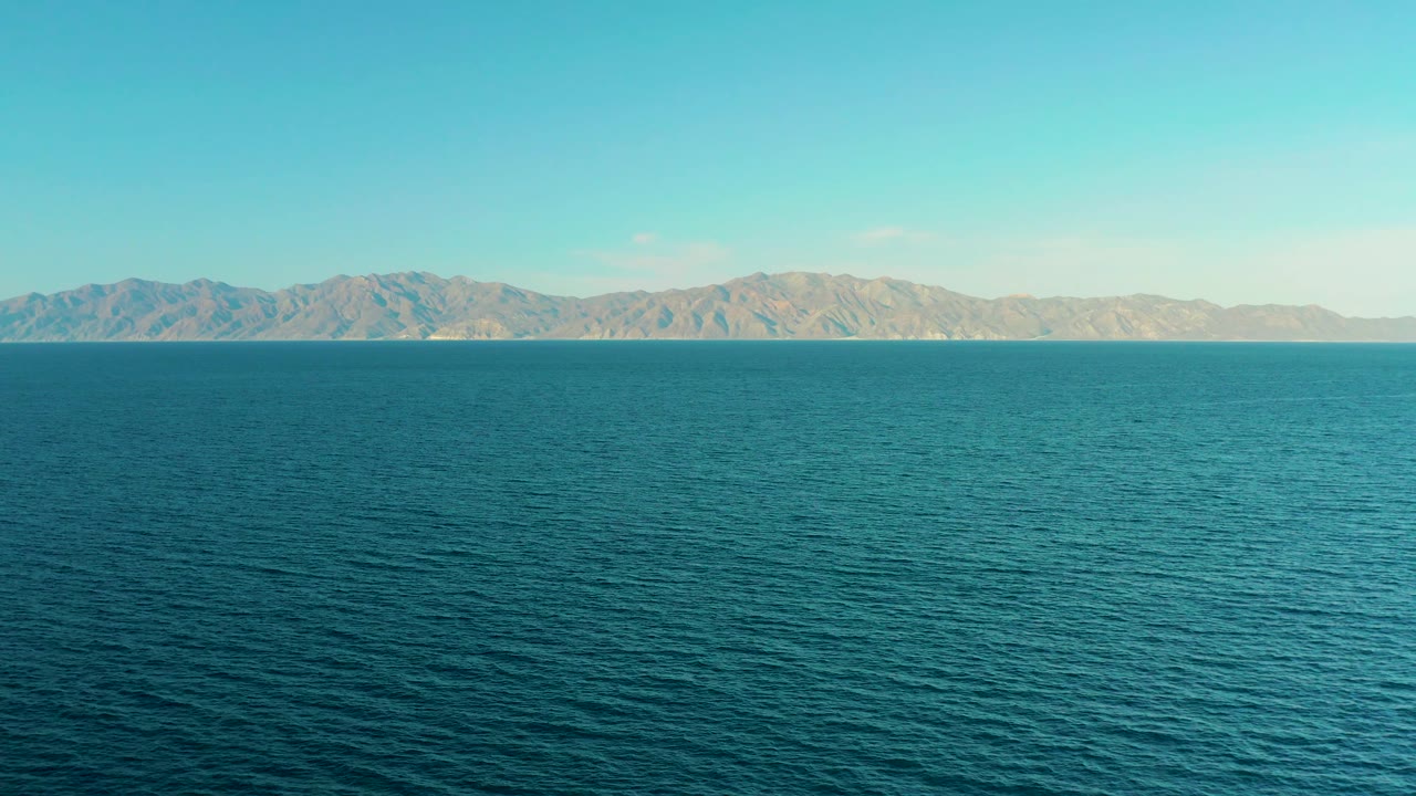 Wide aerial-style view over open blue water toward a low tan mountain range near El Sargento La Ventana, Mexico, with no visible people or vessels.