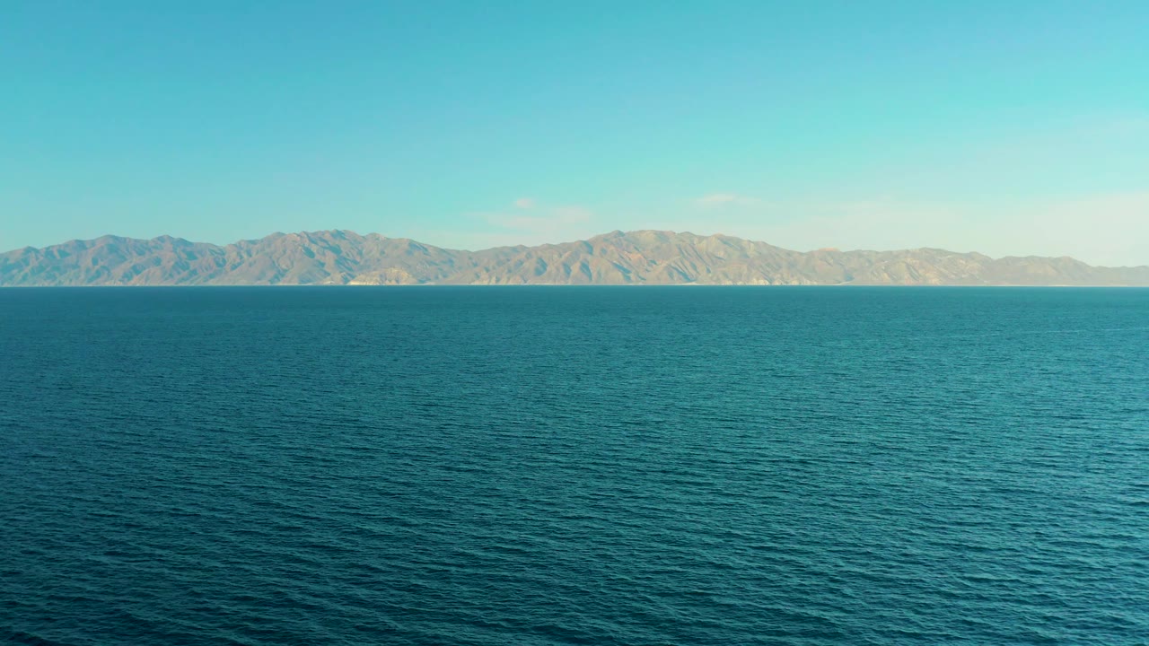 Mid-clip frame with calm blue sea occupying the foreground and tan mountains stretching across the background in El Sargento La Ventana, Mexico, with no visible people.