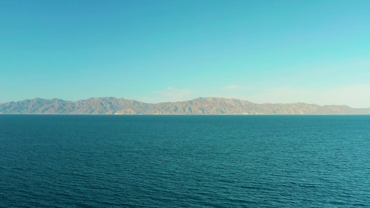 At 0:02, the view holds on rippling turquoise-blue water in front of a rugged tan mountain range near El Sargento La Ventana, Mexico, with clear sky and no visible people.