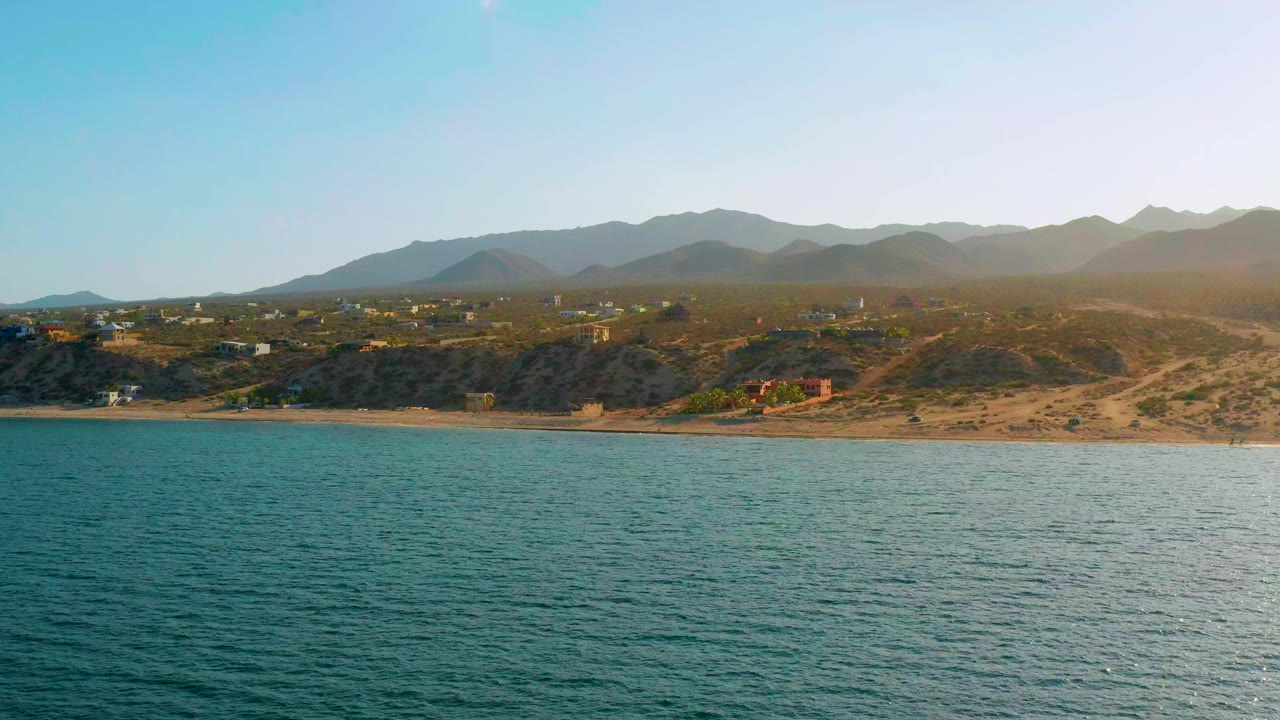 Opening frame of El Sargento La Ventana, Mexico with rippled blue-green water, a narrow sandy beach, scrub-covered dunes, scattered low buildings, hazy mountains, and no visible people.
