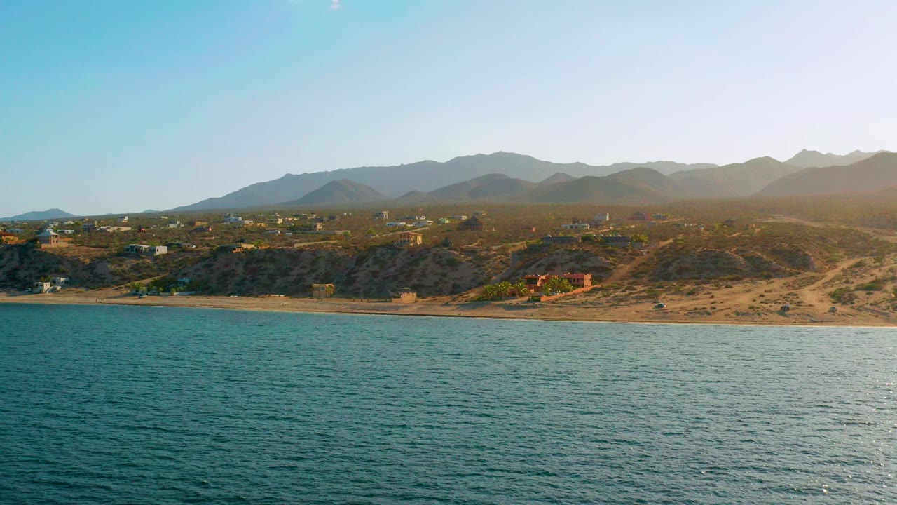 At 0:00, a wide aerial view in El Sargento La Ventana shows blue ocean in front of a sandy shoreline, low coastal hills, scattered buildings, and distant mountains with no visible people.