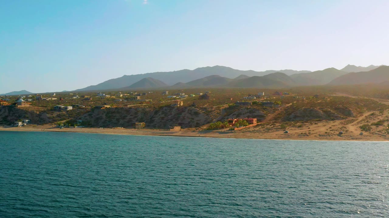 At 0:01, the El Sargento La Ventana coastline remains wide in frame with rippled blue water, a narrow beach, hillside tracks, scattered structures, and hazy mountains with no visible people.