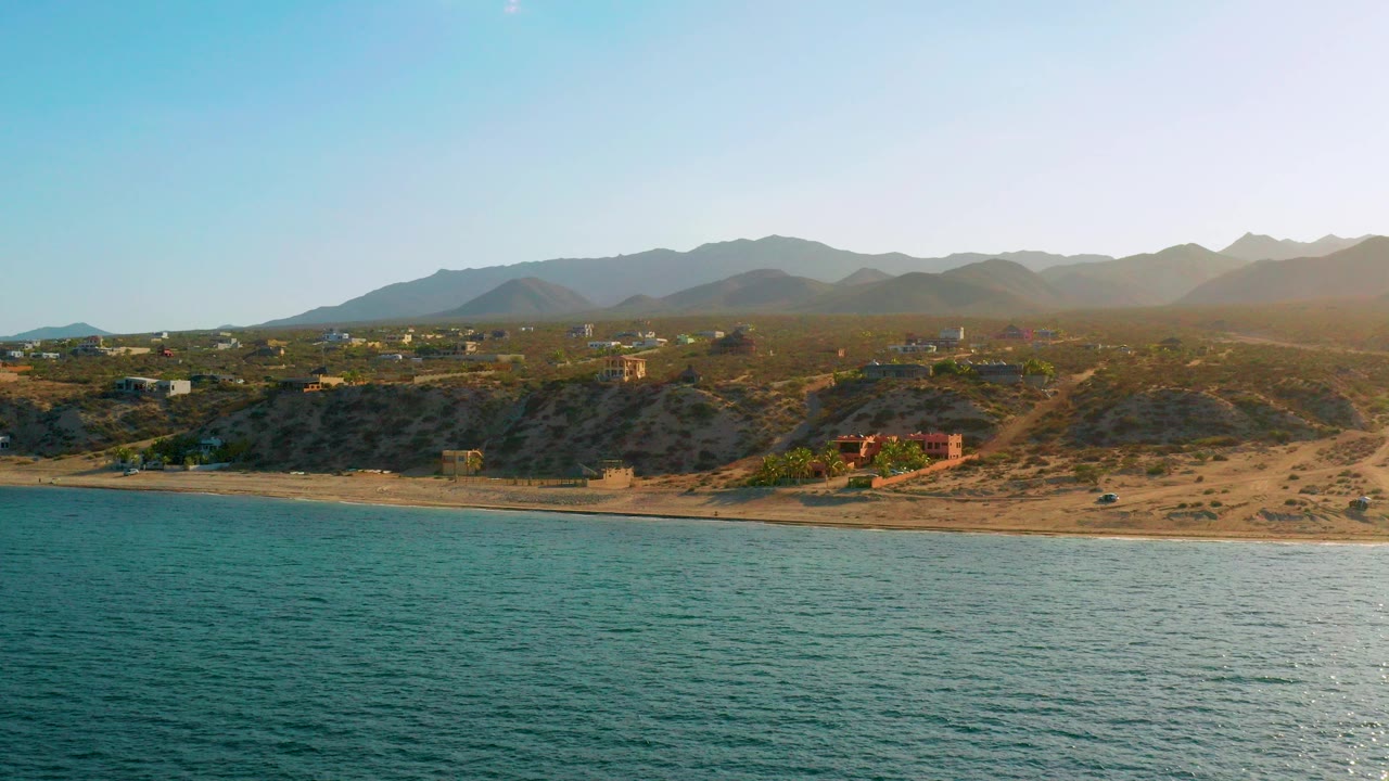 Opening frame of El Sargento La Ventana, Mexico, with blue-green ocean in front, a sandy beach and dry bluffs beyond it, scattered buildings inland, distant mountains, and no visible people.