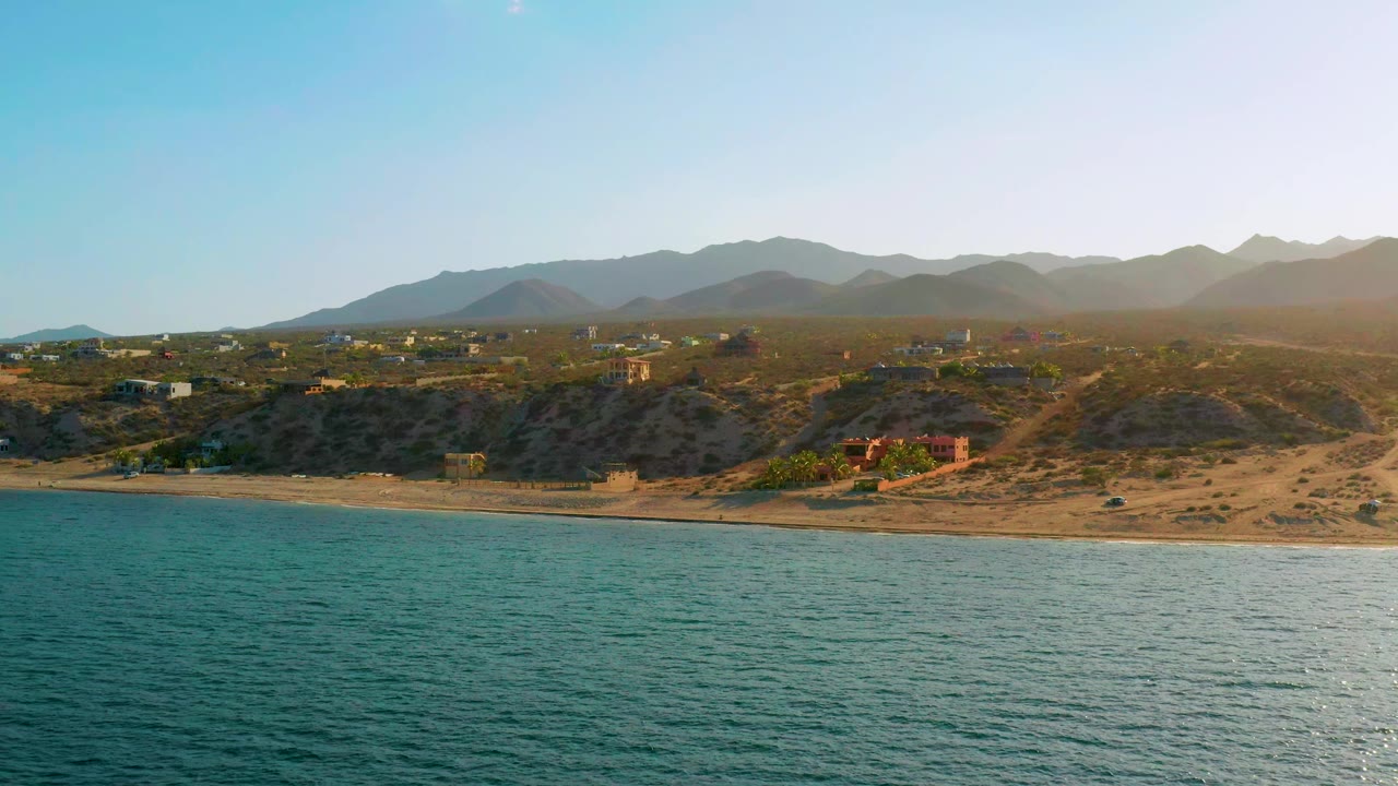 El Sargento La Ventana coastline at 0:01, with textured ocean water across the lower frame, a narrow beach, dry hillside lots, low buildings, layered mountains, and no visible people.