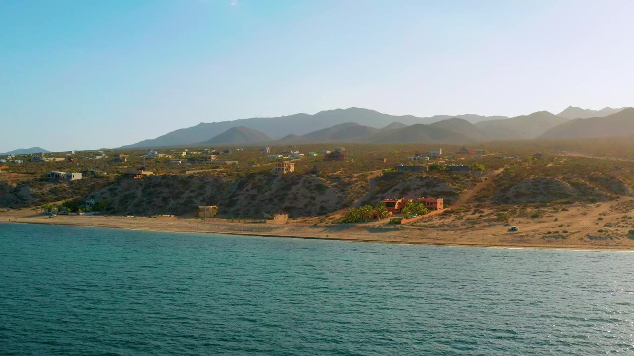 At 0:02 the El Sargento La Ventana view shows rippled ocean in the foreground, a thin surf line, sandy beach, scrubby bluffs, scattered inland structures, hazy mountains, and no visible people.