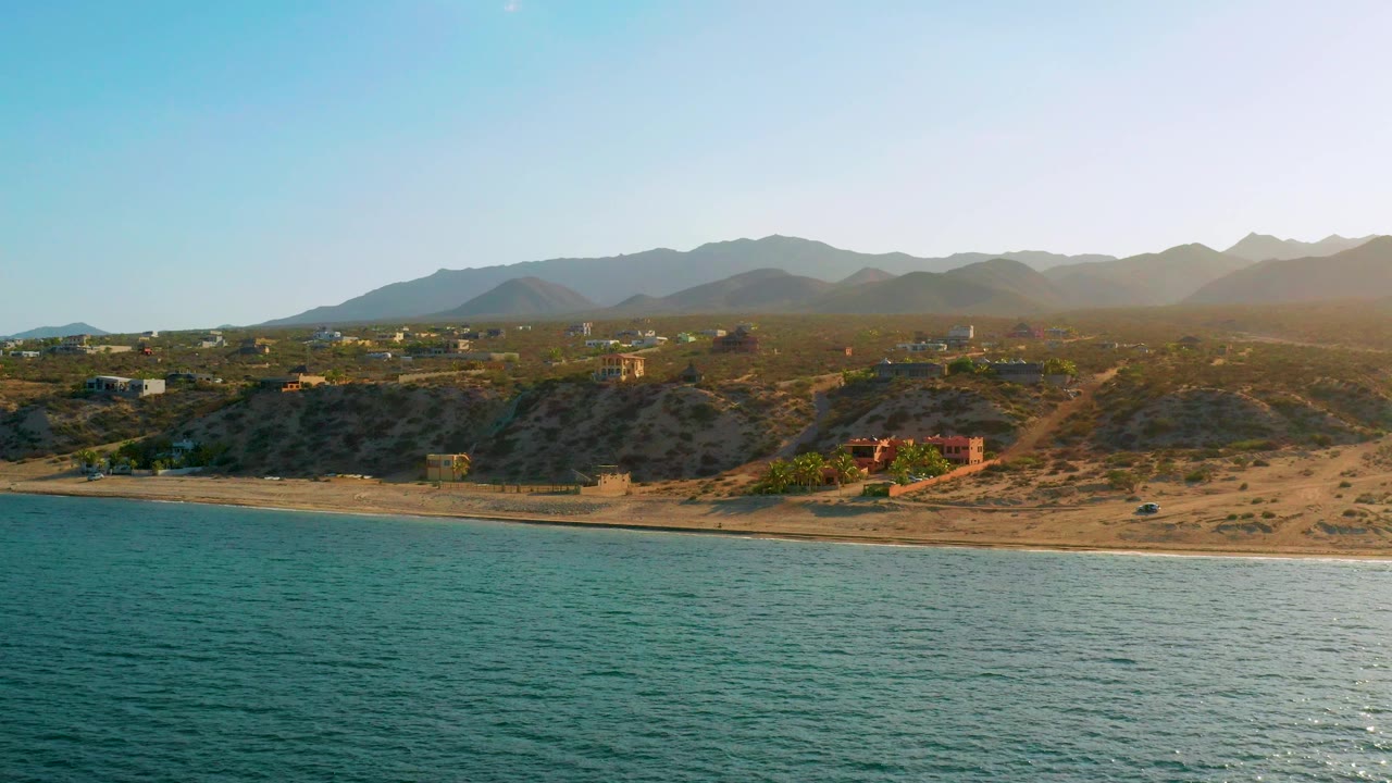 Wide coastal landscape with ocean, beach, arid bluff roads, buildings, and mountains.