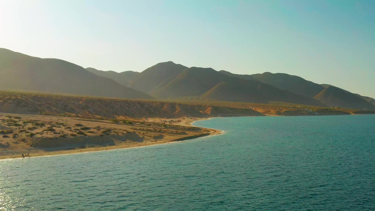 Opening frame over blue water in El Sargento La Ventana, Mexico, with a sandy curve of shore, scrubby hillside, layered mountains, warm light from the left, and no visible people.