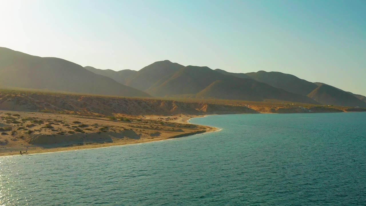 Sunlit desert shoreline and blue water with mountains across the horizon.