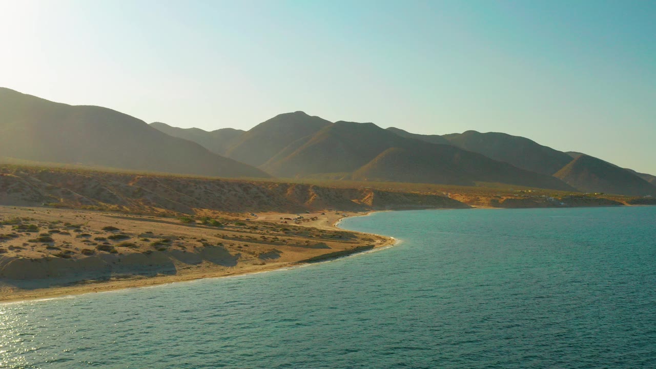 Early frame of El Sargento La Ventana coastline with turquoise water in front, a pale sandy curve, desert scrub, distant mountains, and no visible people.