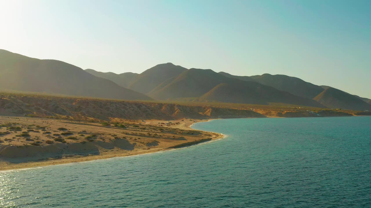 El Sargento La Ventana shoreline with blue water across the foreground, a narrow sand edge, scrub-covered dry land, shadowed mountains, and no visible people.