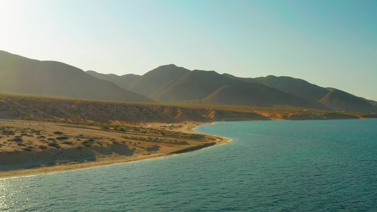 Wide frame from El Sargento La Ventana showing teal sea on the right, a rounded beach arc, ocher desert terrain, dark mountain ridges, and no visible people.