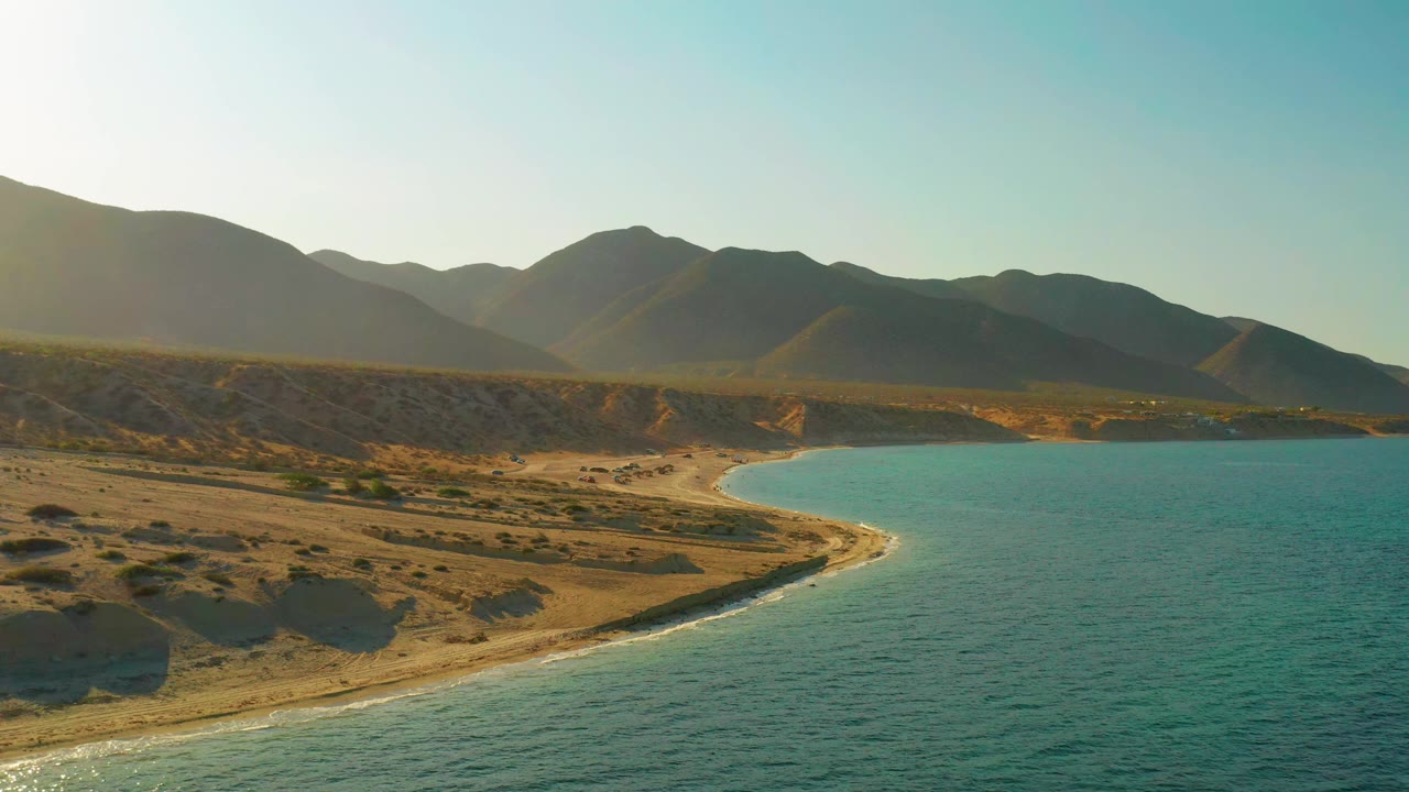 El Sargento La Ventana aerial frame with the cove centered, blue-green water filling the right side, tan shoreline below, hazy mountains behind, and no visible people.