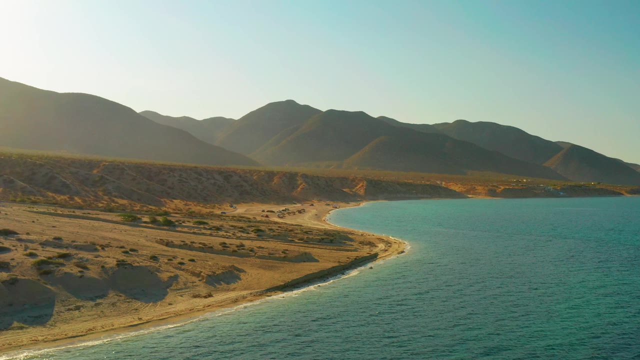 A wide El Sargento La Ventana frame showing desert flats stretching toward a crescent shore, turquoise water at right, shadowed mountains across the horizon, and no visible people.