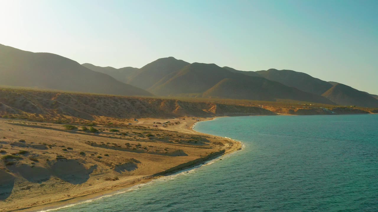 Mid-clip aerial over El Sargento La Ventana with scrub-covered land in the lower frame, small waves tracing the beach, blue water to the right, mountains behind, and no visible people.