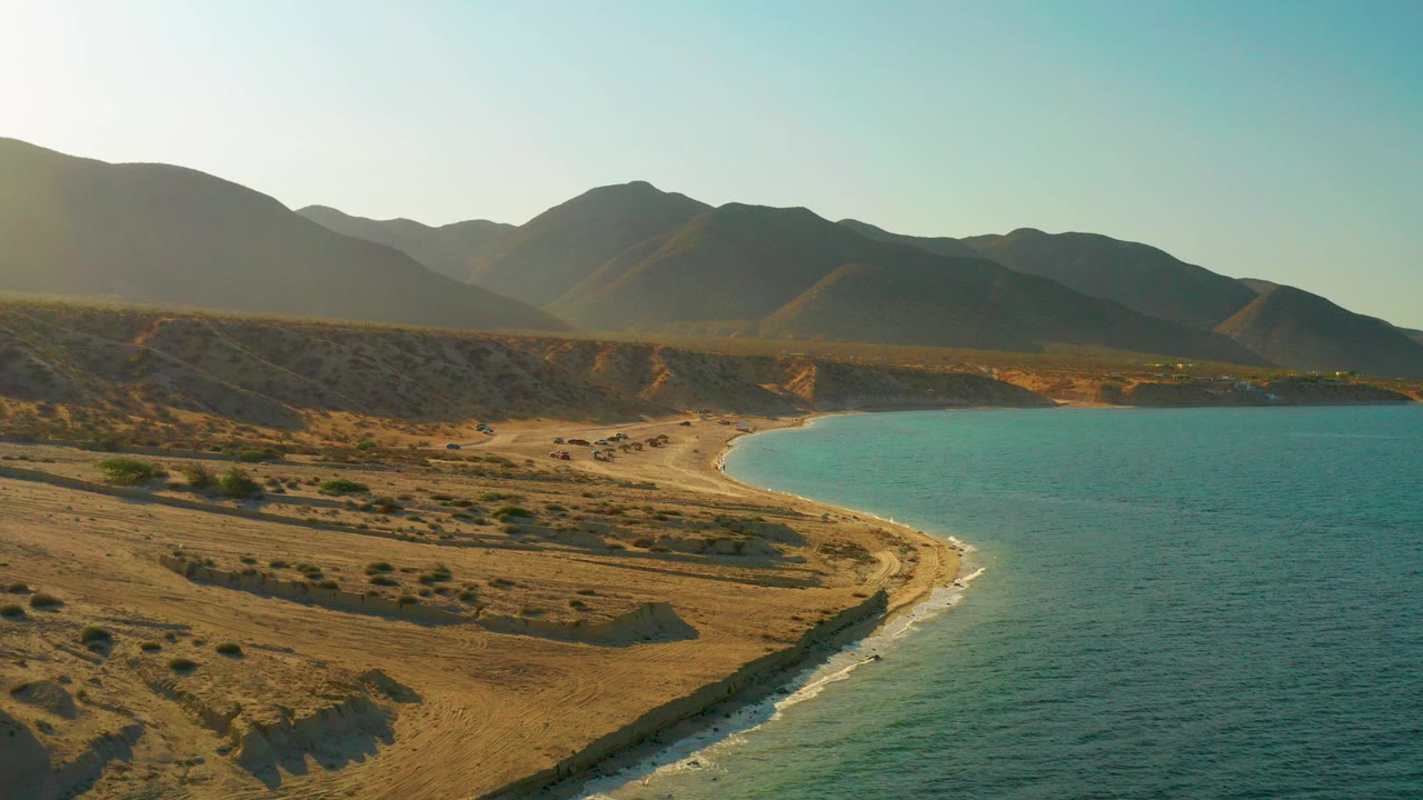 Aerial view over El Sargento La Ventana, Mexico with a curved sandy beach, blue water on the right, dry scrub terrain, distant mountains, and no visible people.