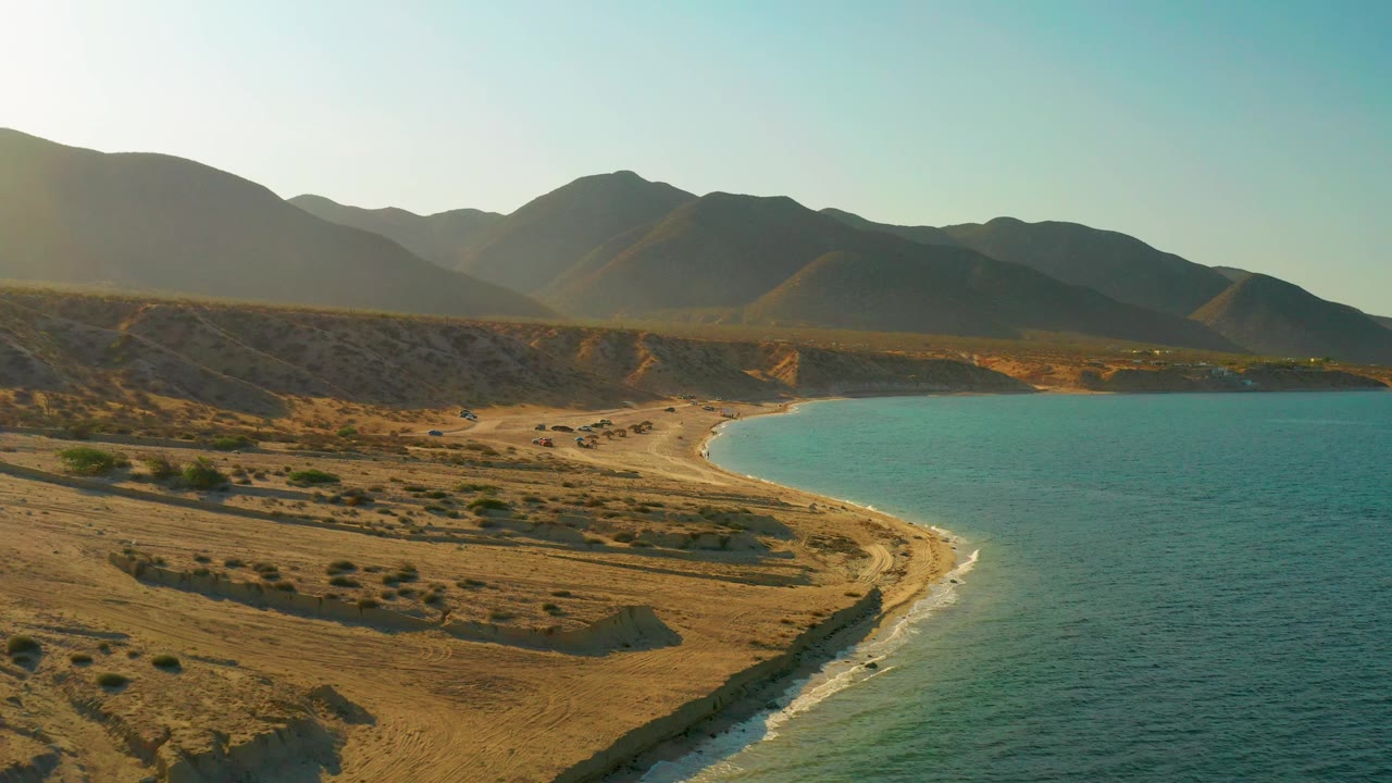 Elevated view over El Sargento La Ventana, Mexico with low waves meeting the sand, open water filling the right side, desert ground left of the beach, and no visible people.