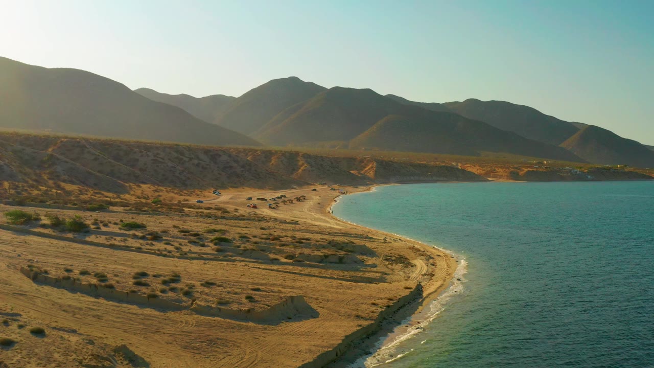 Aerial frame from El Sargento La Ventana, Mexico showing beach access tracks across dry ground, a curved shoreline, blue-green water, mountain shadows, and no visible people.
