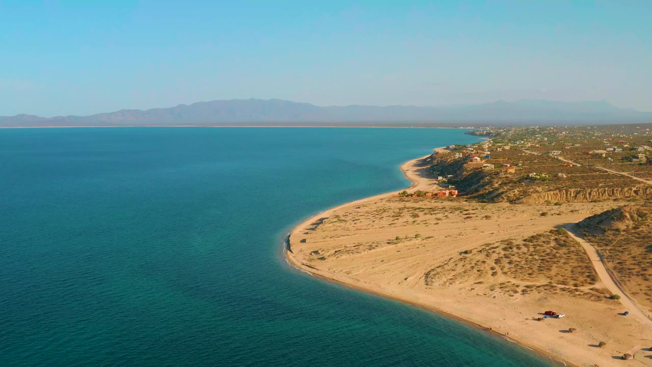 High aerial view in El Sargento La Ventana, Mexico, showing turquoise water beside a rounded sandy point, dry scrub on the dunes, a shoreline road, and no visible people.