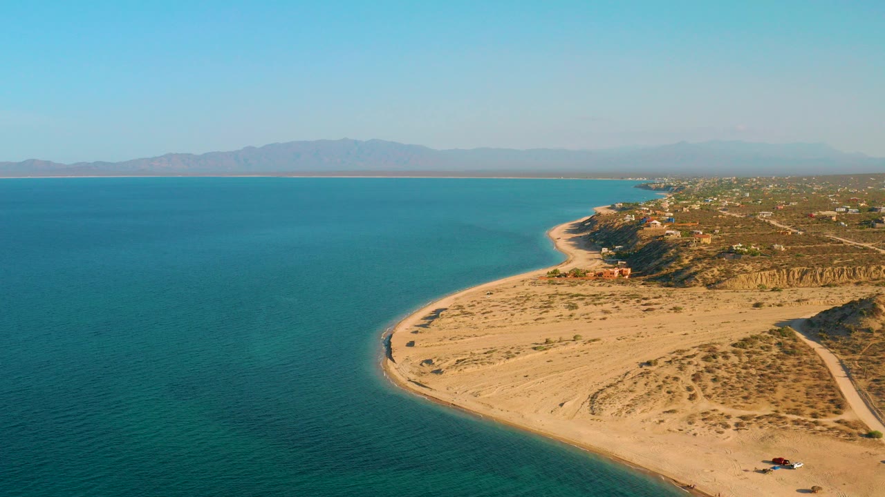 Aerial view of El Sargento La Ventana, Mexico, with turquoise water on the left, a sandy point in the foreground, distant mountains, and no visible people.