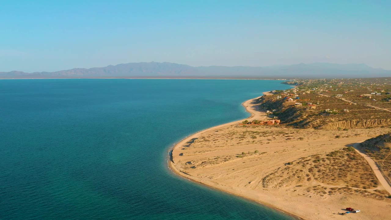 Aerial frame from El Sargento La Ventana, Mexico, with the ocean filling the left side, sand and scrub across the lower right, a coastal community beyond, and no visible people.