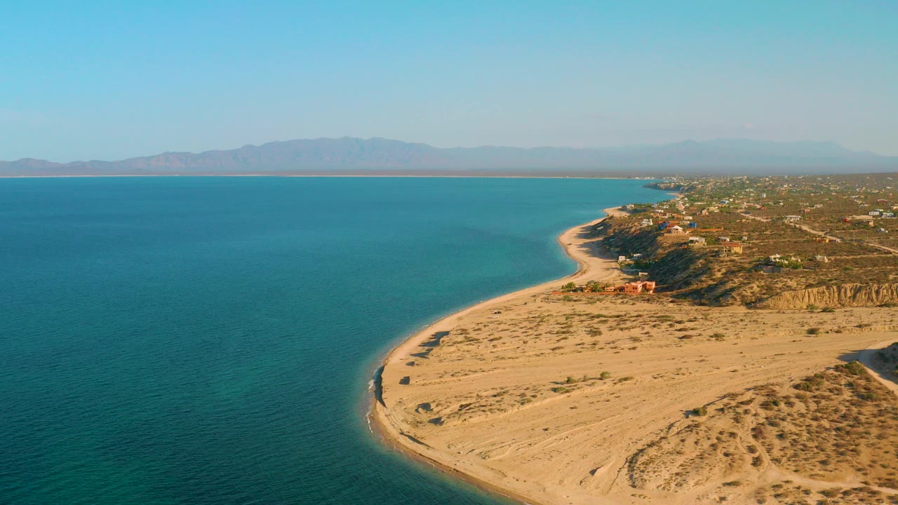 High aerial frame over El Sargento La Ventana, Mexico showing turquoise water, a curving sandy beach, coastal homes, hazy mountains, and no visible people.