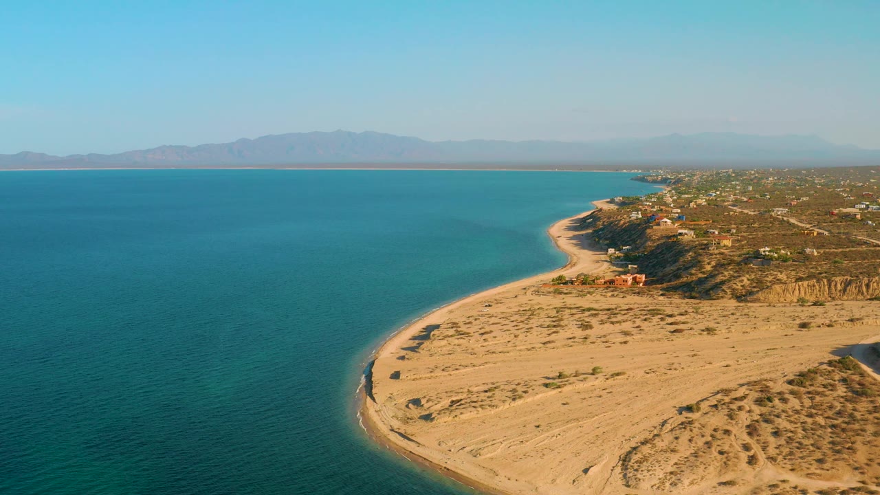Wide drone frame over El Sargento La Ventana, Mexico showing teal water, sunlit sand, beach tracks, bluffside homes, a hazy mountain range, and no visible people.