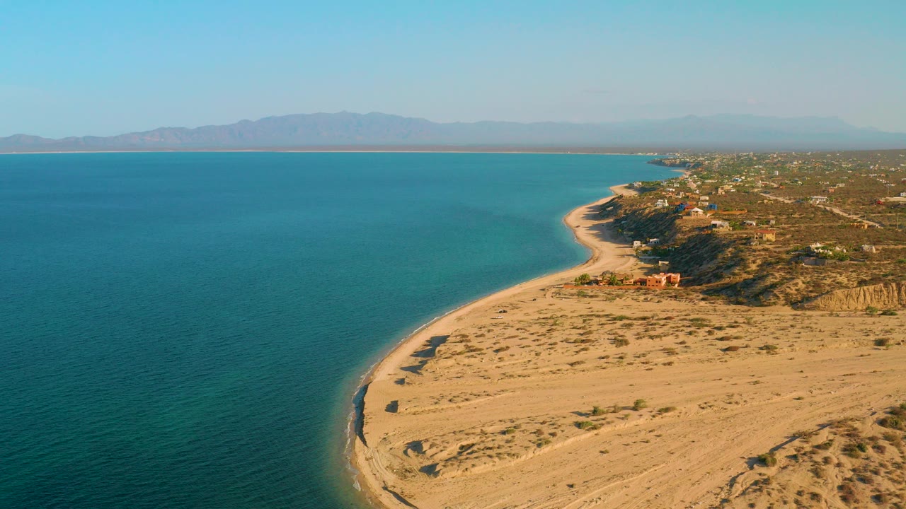 Aerial frame over El Sargento La Ventana, Mexico, with blue ocean on the left, a sandy curved shore on the right, distant mountains, scattered coastal buildings, and no visible people.