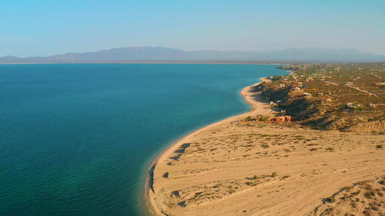 High aerial frame from El Sargento La Ventana, Mexico, with broad blue sea, pale sand, a curving shoreline, low coastal buildings, distant mountains, and no visible people.