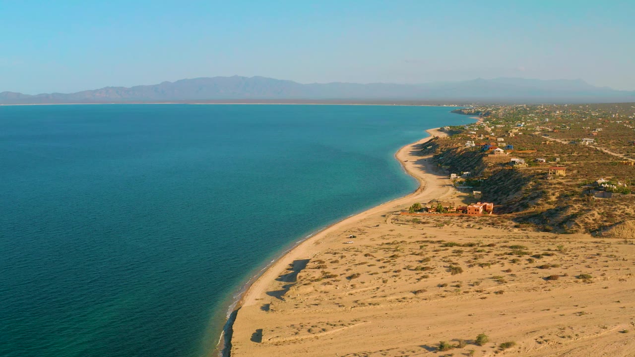 Aerial view one second into the clip shows the El Sargento La Ventana shoreline with calm blue water, pale sand, scrub-covered dunes, scattered coastal structures, hazy mountains, and no visible people.