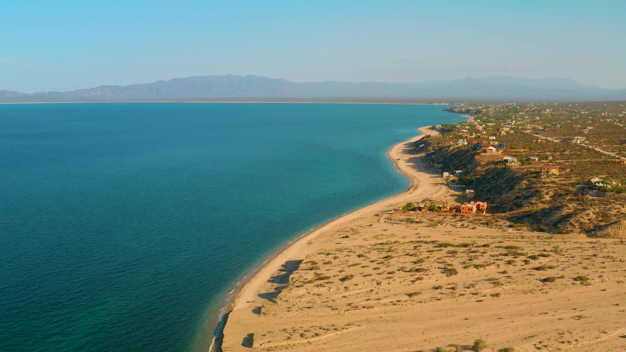 High aerial frame of El Sargento La Ventana, Mexico, with turquoise sea taking most of the left frame, a narrow sandy beach bending toward the town, dry terrain to the right, distant mountains, and no visible people.