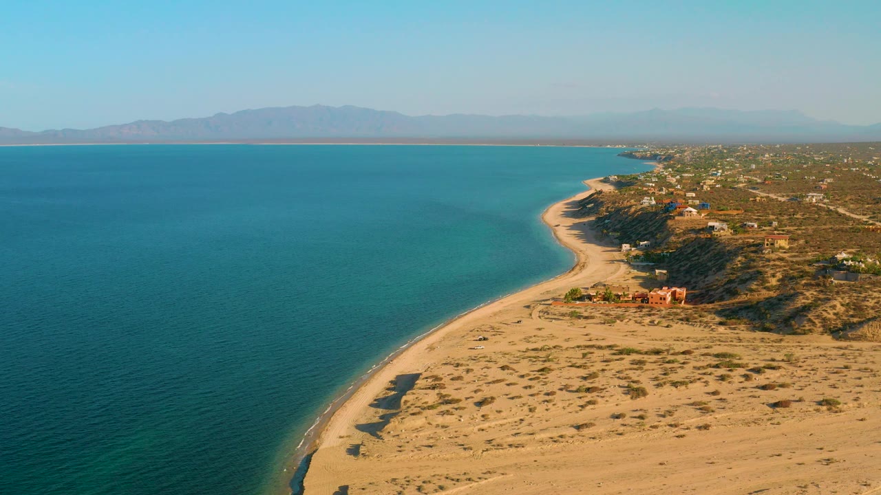 Mid-clip aerial view over El Sargento La Ventana shows blue water on the left, a sunlit beach and dunes through the center, scattered buildings on the right, a hazy mountain line beyond the bay, and no visible people.