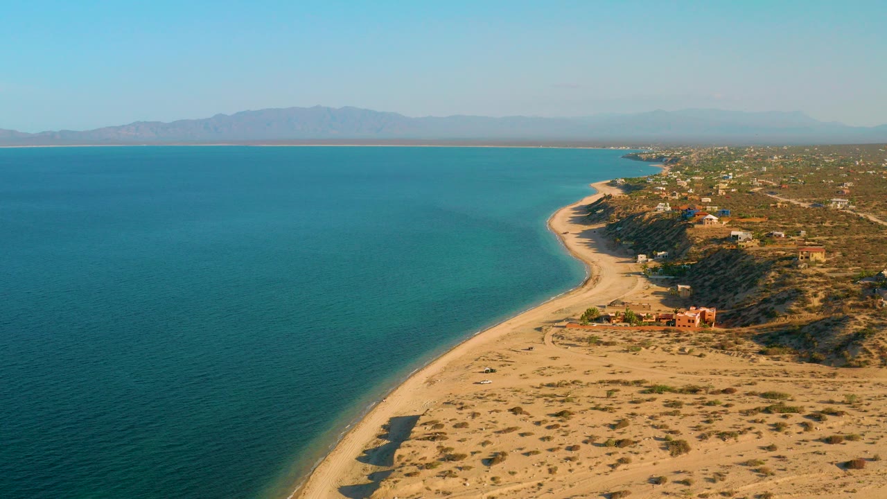 Aerial view over El Sargento La Ventana, Mexico, with blue ocean on the left, a curved sandy beach below, coastal buildings to the right, distant mountains, and no visible people.
