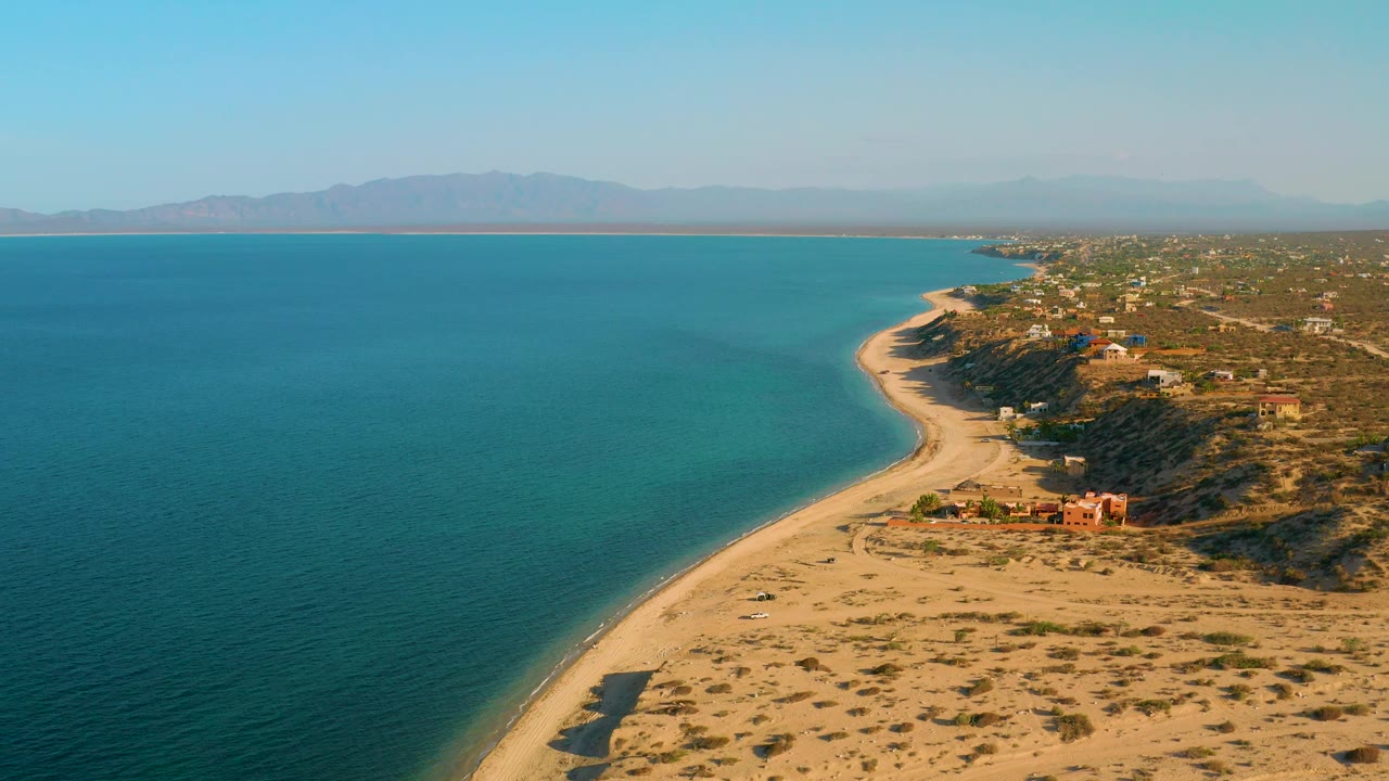 Wide aerial frame of El Sargento La Ventana, Mexico, with broad blue water, a narrow sandy shoreline, low inland structures, dry scrub terrain, and no visible people.