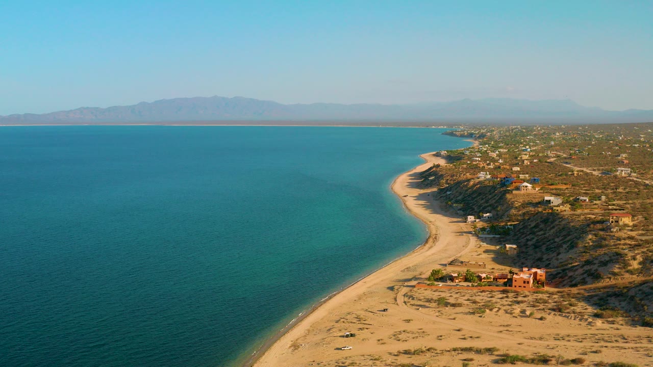 Opening aerial frame over El Sargento La Ventana, Mexico, with turquoise water on the left, sandy beach curving along the shore, scattered homes to the right, distant mountains, and no visible people.
