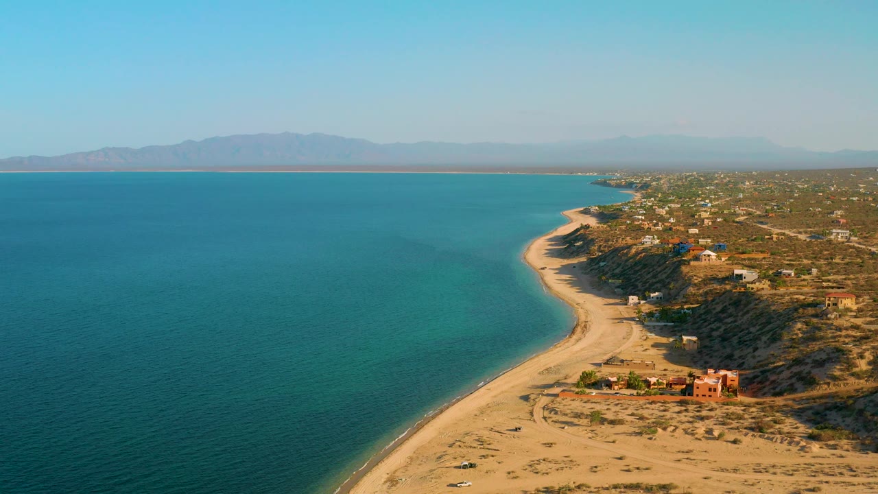 High aerial view over El Sargento La Ventana, Mexico, with more open ocean filling the left side, pale sand and a few beachfront structures on the right, distant mountains, and no visible people.
