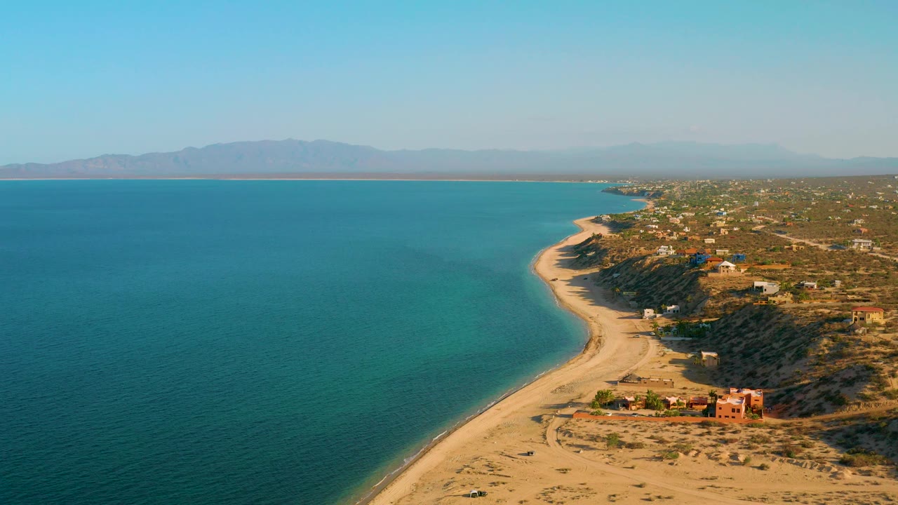 Aerial shoreline with calm sea, sandy beach, scrubland, and distant mountains.