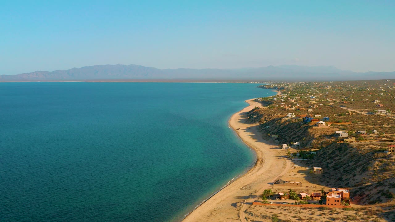 Opening aerial frame over El Sargento La Ventana, Mexico, with turquoise water on the left, a pale beach curving below, scattered bluff-top buildings to the right, distant mountains, and no visible people.