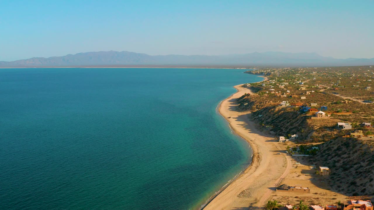Aerial frame over El Sargento La Ventana, Mexico showing turquoise water, a pale curving beach, scattered coastal buildings, distant mountains, and no visible people.