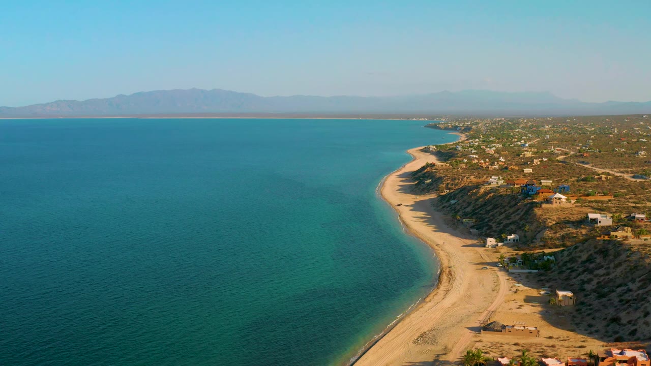 Wide drone view of blue-green water and a sandy shoreline with houses inland.