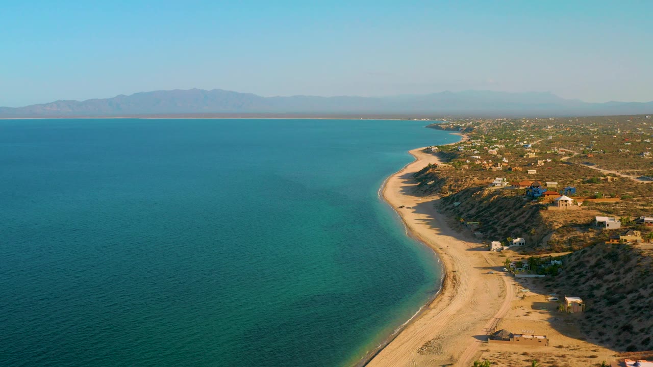 Aerial view of El Sargento La Ventana, Mexico with open turquoise water, pale beach, scrub-covered coastal land, small buildings, distant mountain silhouettes, and no visible people.