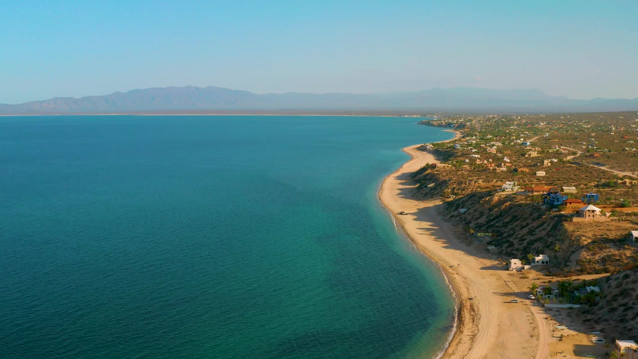 High aerial frame over El Sargento La Ventana showing clear water on the left, a narrow sandy beach at center, scrubby hillside with scattered houses on the right, distant mountains, and no visible people.
